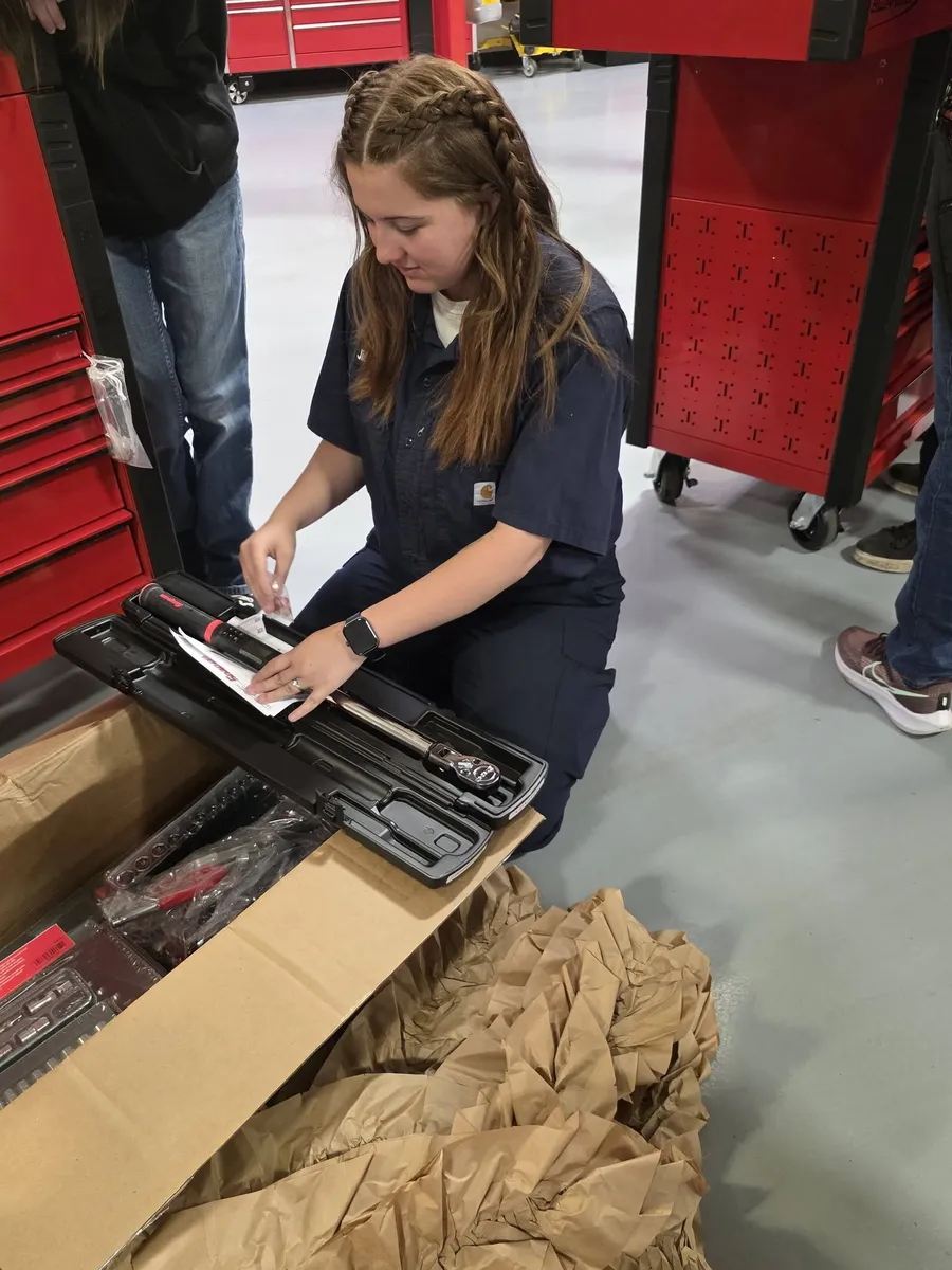 Jillian in a navy work shirt, kneeling to unpack the new tools from her scholarship, a red tool chest behind her