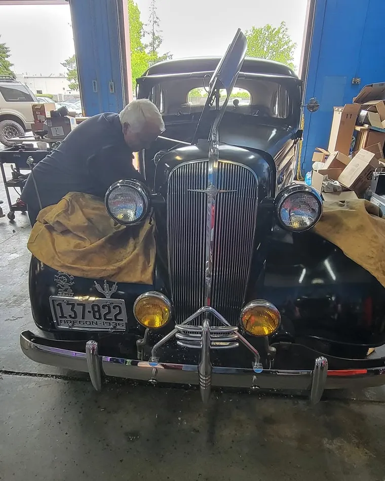 Russ leaning into the engine bay of a black 1936 Chevy inside the shop, the bay door open behind him